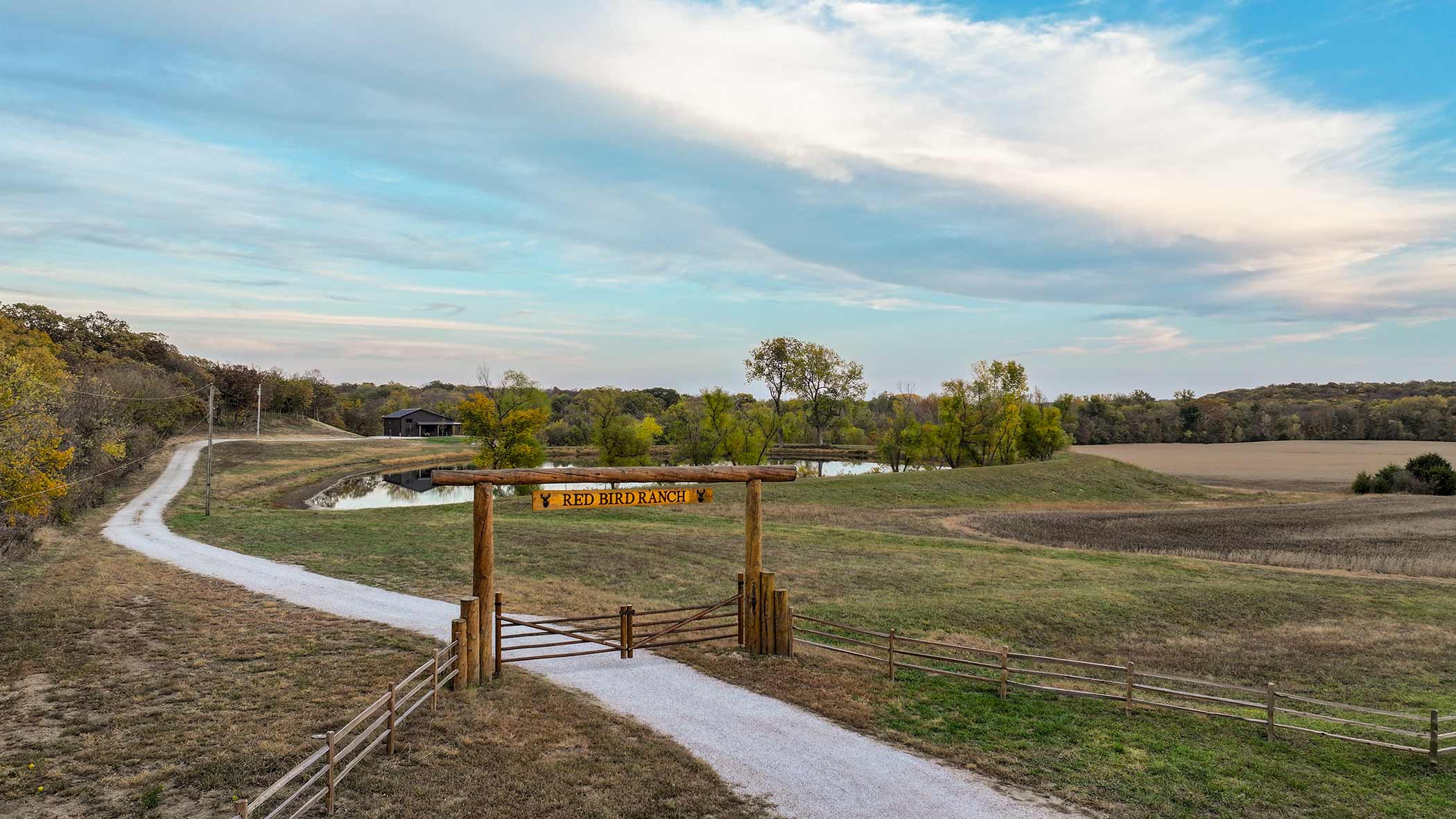 Aerial view of Red Bird Ranch entrance gate with gravel drive, lake, and lodge in autumn