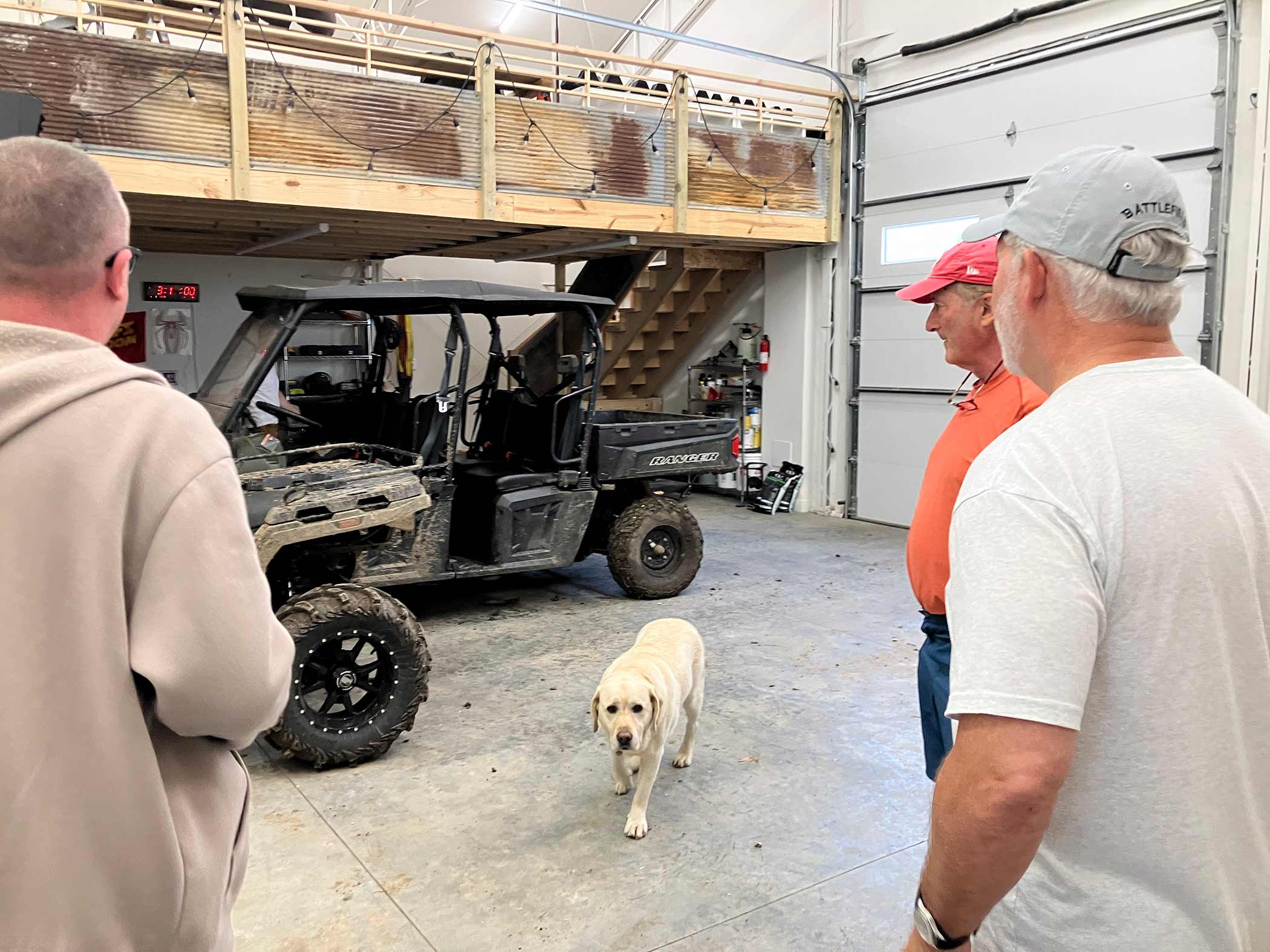 Two guests and the ranch yellow Lab in the gear bay — muddy Polaris Ranger UTV, mezzanine loft above, bay doors open