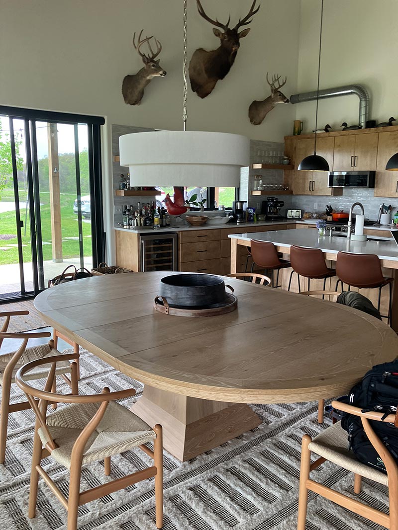 Red Bird Ranch dining room — round wood table with wishbone chairs, three trophy mounts on a high wall, wine fridge and glass doors