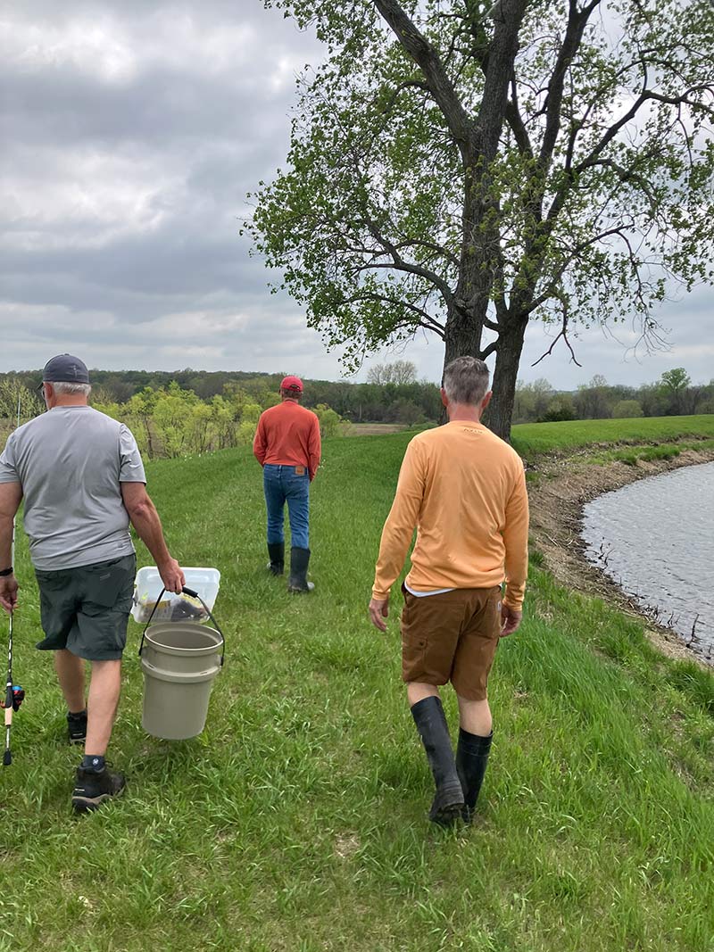 Three guests walking the lake bank at Red Bird Ranch — fishing rods, rubber boots, dramatic sky