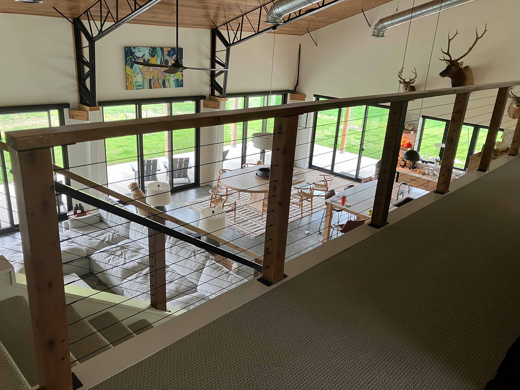 Mezzanine aerial view of the Red Bird Ranch great room — vaulted wood trusses, floor-to-ceiling glass walls, stone fireplace, and the Kansas lawn beyond